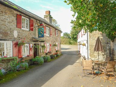 A stone building with red shutters and flower boxes along a narrow street with outdoor table and chairs under a tree at 1 Gabberwell Cottages in Kingston