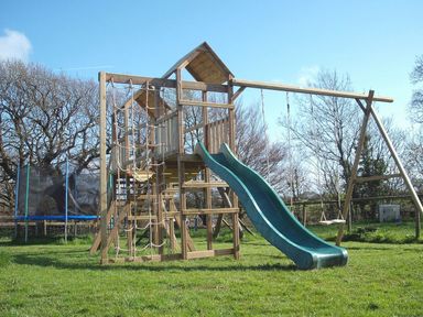 A wooden play structure with a slide swings and climbing ropes in a grassy yard at Yellowland Farm in Holsworthy