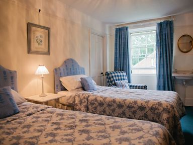 A bedroom with two single beds beside a window with patterned blue curtains and a checked armchair at Parnacott in Holsworthy
