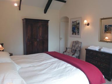 A bedroom with a bed covered by white sheets and red blanket a dark wooden wardrobe a patterned armchair and a dresser with folded towels at Higher Tumrose in Blisland