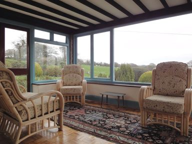 A seating area with three cushioned armchairs and a small table near large windows overlooking a garden at Higher Tumrose in Blisland