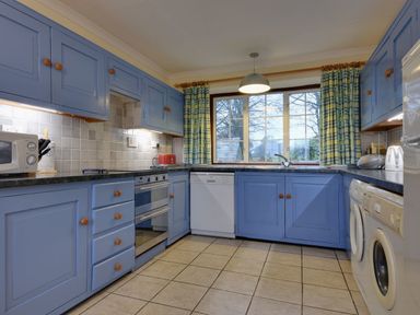 A kitchen with blue cabinets a dishwasher oven washing machine and a window with checkered curtains at Higher Tumrose in Blisland