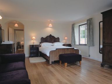 A bedroom with a wooden bed and bedside tables with lamps a window with curtains a dark ottoman and a wooden wardrobe at Higher Tumrose in Blisland