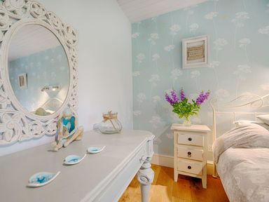 A bedroom corner with a white wooden table holding a mirror statue and dishes beside a nightstand with flowers and a bed in the room at Grandpa Dicksons in Little Petherick
