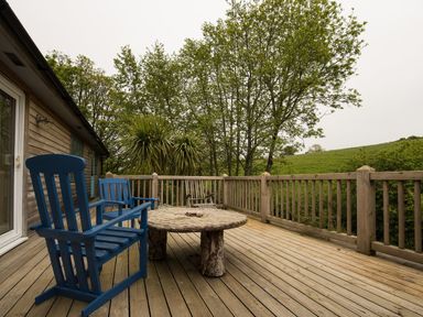 An outdoor wooden deck with blue chairs and a round wooden table surrounded by trees at Grandpa Dickson's in Little Petherick