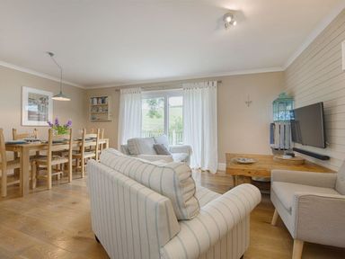 A living room and dining area with striped sofas a wooden dining table and chairs a wall mounted TV and white curtains at Grandpa Dicksons in Little Petherick