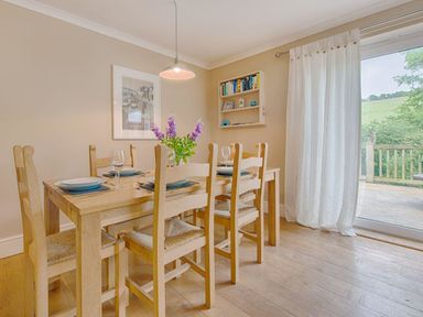 A dining room with wooden table and chairs set with plates and glasses near a sliding door with white curtains at Grandpa Dickson's in Little Petherick