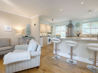 A living room with armchairs and a sideboard adjacent to a kitchen with a breakfast bar and stools at Grandpa Dickson's in Little Petherick