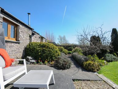A stone wall with a window and a chimney next to a garden path with bushes and a tree and outdoor seating with a table and chair at Swallow Barn in St Kew
