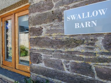 A stone exterior wall with a wooden framed window and a sign that reads Swallow Barn at Swallow Barn in St Kew