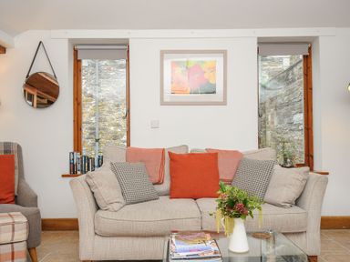A living room with a beige sofa and cushions between two windows with a round mirror on the wall at Swallow Barn in St Kew