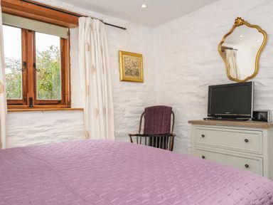 A bedroom with a purple bedspread a wooden chair with a blanket a dresser with a television and mirror at Swallow Barn in St Kew