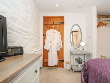 A bedroom with a wooden door with white robe hanging a bedside table with a mirror and flowers at Swallow Barn in St Kew