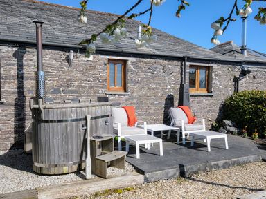 An outdoor seating area with two chairs, two tables, and a wooden hot tub against a stone building at Swallow Barn in St Kew