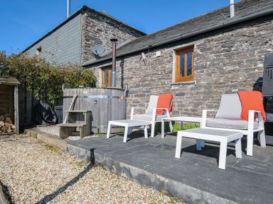 An outdoor patio area with white chairs and tables and a wooden hot tub next to a stone building at Swallow Barn in St Kew
