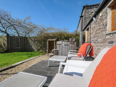 An outdoor seating area with lounge chairs, a small table, a wooden hot tub, green grass, and a wooden fence at Swallow Barn in St Kew