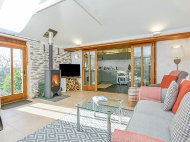 A living room with a glass coffee table a fireplace and a sofa facing a kitchen at Swallow Barn in St Kew