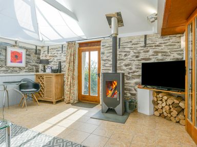 A room with a wood burning stove a stack of firewood a television a wooden cabinet a black chair and a table at Swallow Barn in St Kew