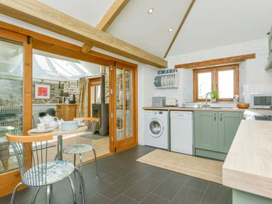 A kitchen area with green cabinets, washing machine, dishwasher, round table with two chairs and wooden beams at Swallow Barn in St Kew