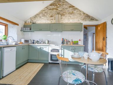 A kitchen with green cabinets a wooden door a small round table with chairs and kitchen appliances at Swallow Barn in St Kew