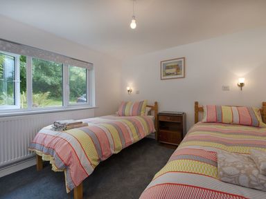 A bedroom with two single beds with striped bedding a wooden nightstand between the beds and a window with a view of greenery at Salty Cottage Morwenstow