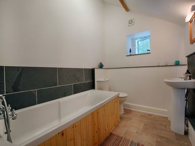 A bathroom with a bathtub wooden panel toilet pedestal sink small window and tiled floor at Salty Cottage in Morwenstow