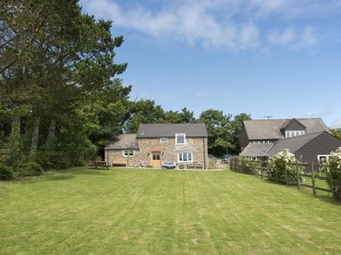 A grassy garden with a stone house and trees in the background at Salty Cottage in Morwenstow