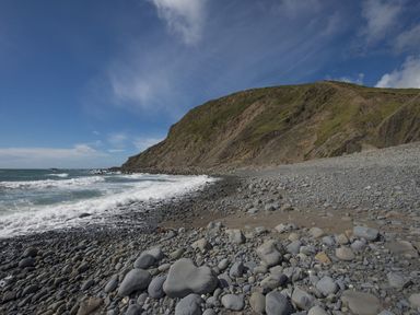 A rocky beach with waves hitting the shore and a large cliff in the background at Salty Cottage in Morwenstow