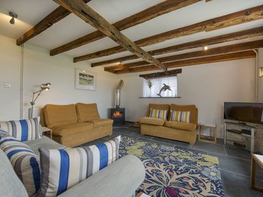 A living room with wooden ceiling beams armchairs a patterned rug a small stove and a television at Salty Cottage in Morwenstow