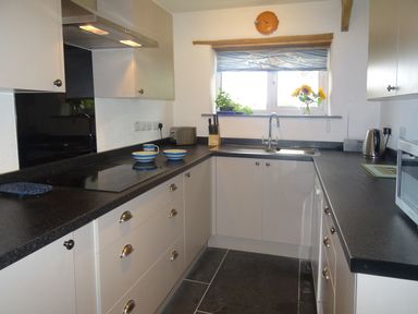 A kitchen with black countertops white cabinets a stove sink window with blinds and a kettle at Salty Cottage in Morwenstow