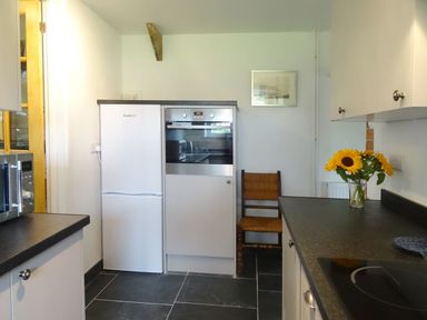 A kitchen with white cabinets black countertops a microwave a built in oven a refrigerator a wooden chair and a vase of sunflowers on the counter at Salty Cottage in Morwenstow