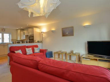 A living room with red sofas two wooden side tables and a television next to a kitchen at The Loft in Bude