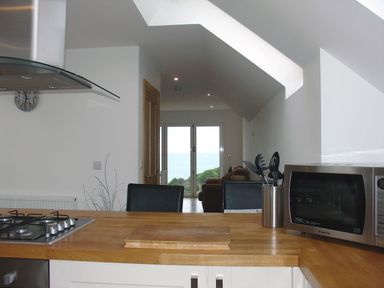 A kitchen with a gas stove and wooden countertop looking out to a living area with black chairs and glass doors showing a view of the sea at Seagulls Perch in Mawgan Porth