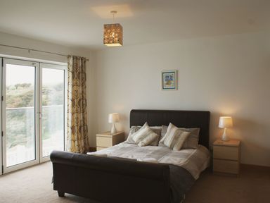 A bedroom with a bed with pillows two bedside tables with lamps and a glass door with curtains at Seagulls Perch in Mawgan Porth