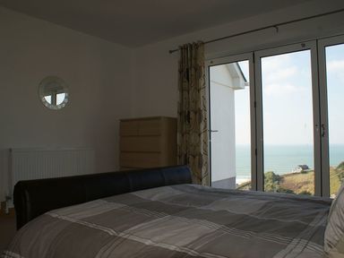 A bedroom with a bed a dresser a round mirror and large glass doors overlooking the sea at Seagulls Perch in Mawgan Porth
