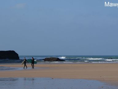 Two surfers walking on a sandy beach with surfboards near the ocean at Seagulls Perch in Mawgan Porth