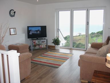 A living room with brown sofas a striped rug a television and large glass doors showing a hill and sea view at Seagulls Perch in Mawgan Porth