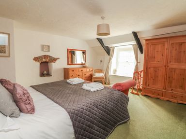 A bedroom with a bed covered with a quilt and pillows a wooden dresser with a mirror a wicker chair a window with curtains a wooden wardrobe and a red upholstered bench at Cousham Cottage Kingsand And Cawsand