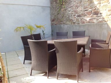 An outdoor patio area with a rectangular glass table and eight wicker chairs surrounded by stone and plaster walls with a small palm plant at Nirvana Kingsand And Cawsand
