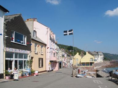 A seaside street with shops, houses, a flag, and people sitting near the shore at Nirvana in Kingsand And Cawsand