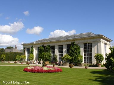 A white building with large windows surrounded by green plants and a flower bed on a lawn at Mount Edgcumbe