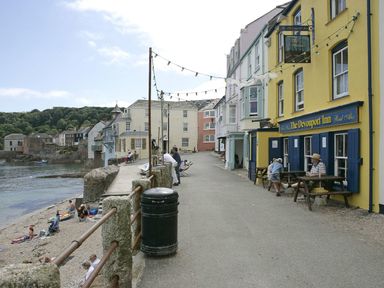 A seaside street with colorful buildings and people sitting outside The Devonport Inn near a beach with people and water at Nirvana in Kingsand And Cawsand