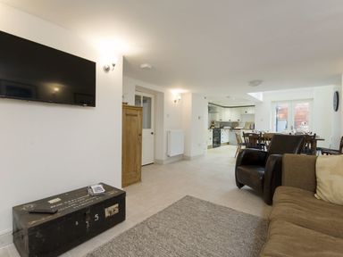 A living room with a wall-mounted television a black trunk a brown sofa and chair a dining area and kitchen in the background at Nirvana Kingsand And Cawsand