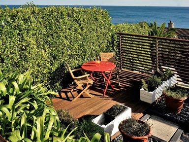 An outdoor wooden deck with a red round table and two wooden chairs surrounded by plants and overlooking the sea at Rock Rose in Downderry