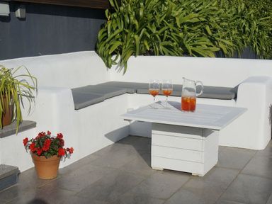 An outdoor seating area with a white built-in bench with gray cushions a white square table with a pitcher and two glasses of orange drink a potted plant with red flowers and green leaves at Rock Rose in Downderry