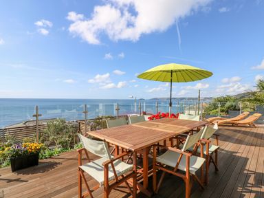An outdoor wooden deck with dining table and chairs a yellow umbrella and two wooden loungers overlooking the sea at Rock Rose in Downderry