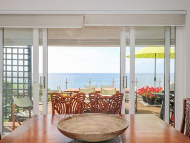 A dining room with wooden chairs and a round wicker bowl on a table overlooking a patio with chairs and a yellow umbrella by the sea at Rock Rose in Downderry