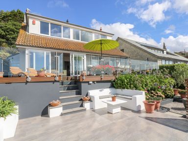 An outdoor patio area with white seating and potted plants in front of a house with glass doors and a green umbrella at Rock Rose in Downderry