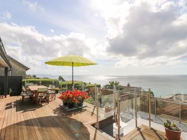 A wooden deck with a table and chairs under a green umbrella and potted flowers overlooking the sea at Rock Rose in Downderry