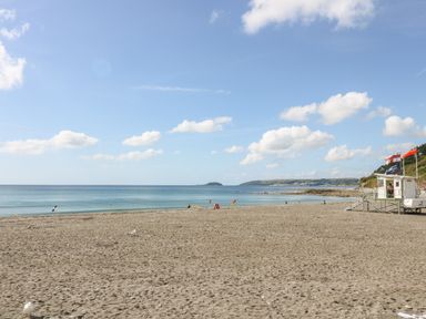 A sandy beach with a lifeguard hut and flags near the shoreline with a calm sea and partly cloudy sky at Rock Rose in Downderry